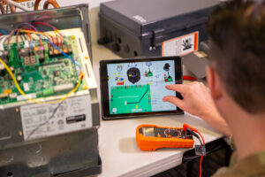A US military member manipulates 3D images on a tablet open to the BILT app. To his left sits electronic components with multicolored wiring. The image on the tablet shows instructions for how to wire the parts correctly.