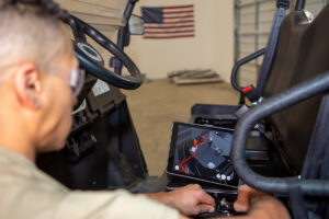 Over the shoulder view of a young man in the US military uses the BILT app on a touchscreen. Vehicle parts are in the background.