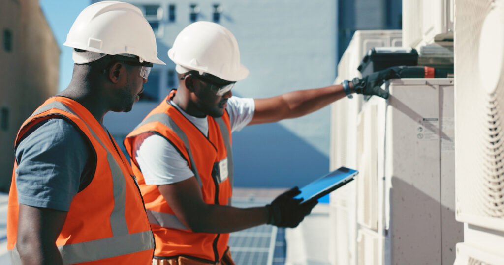 Two men in field service training use interactive instructions to install hvac equipment. One man in the background holds a tablet in his right hand.