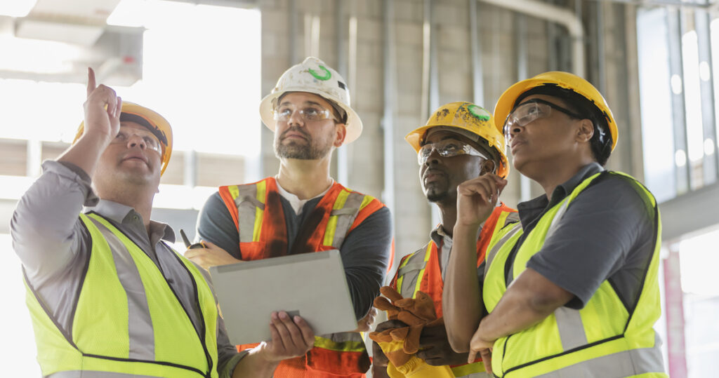 A multi-ethnic group of four construction workers wearing hardhats and safety vests in a training program inside the structure being built. A man is holding a digital tablet and pointing up toward the ceiling.